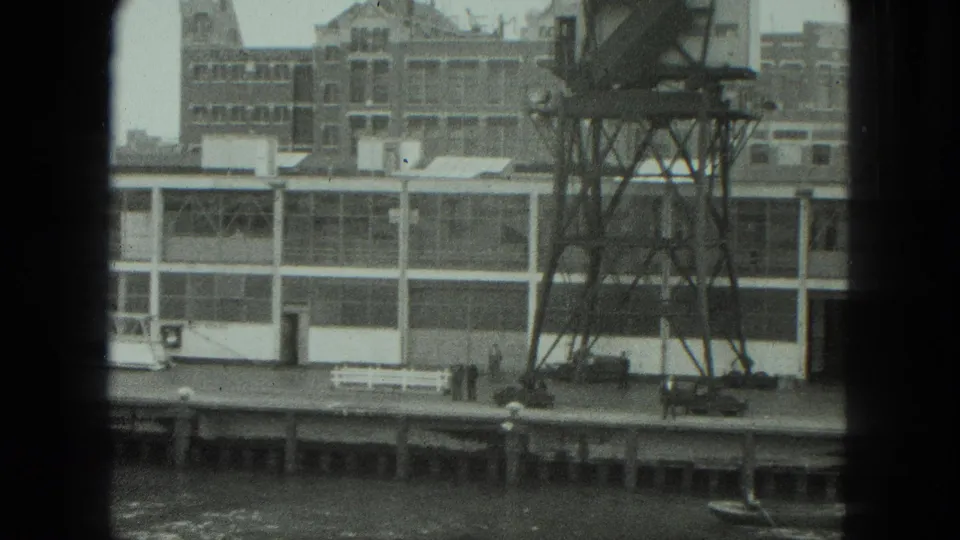 Black-and-white 8mm frame of the Venice waterfront showing the factory facade, tower structure, dock edge, and distant people near the pier.