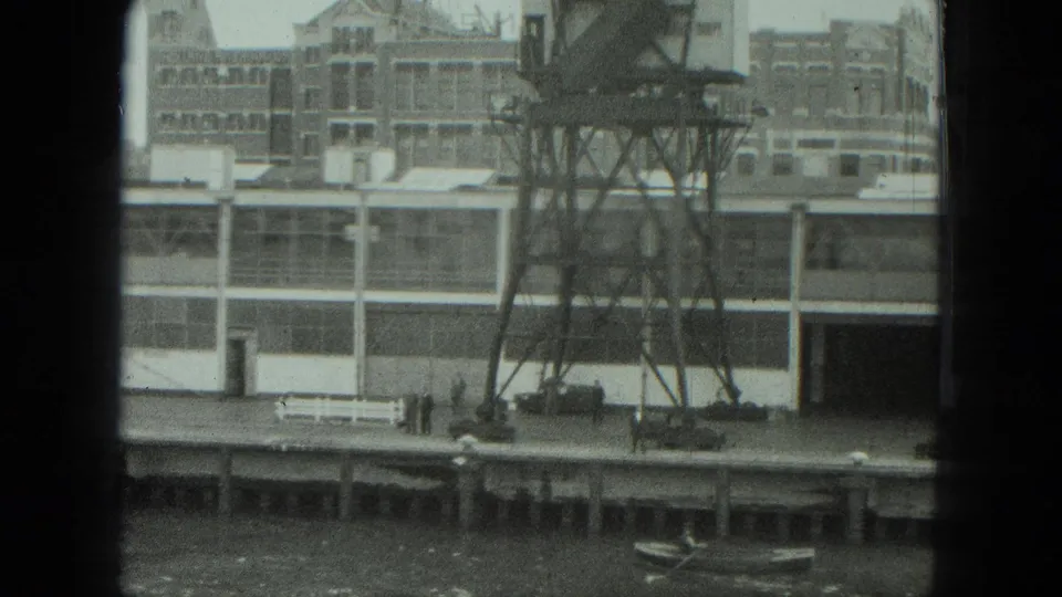 1975 Venice home-movie frame showing an industrial building along the river, a metal tower, dockside bench, vehicle, and a small boat at lower right.