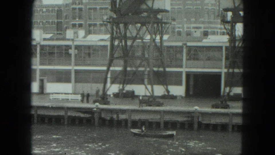 Black-and-white 8mm view of a Venice industrial waterfront with a metal tower, factory facade, dockside figures, parked vehicles, and a small boat.