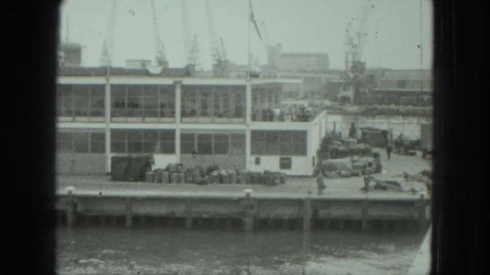 1975 Venice waterfront scene with a glass-fronted dock building, gathered people, cranes, cars, and water in the foreground.