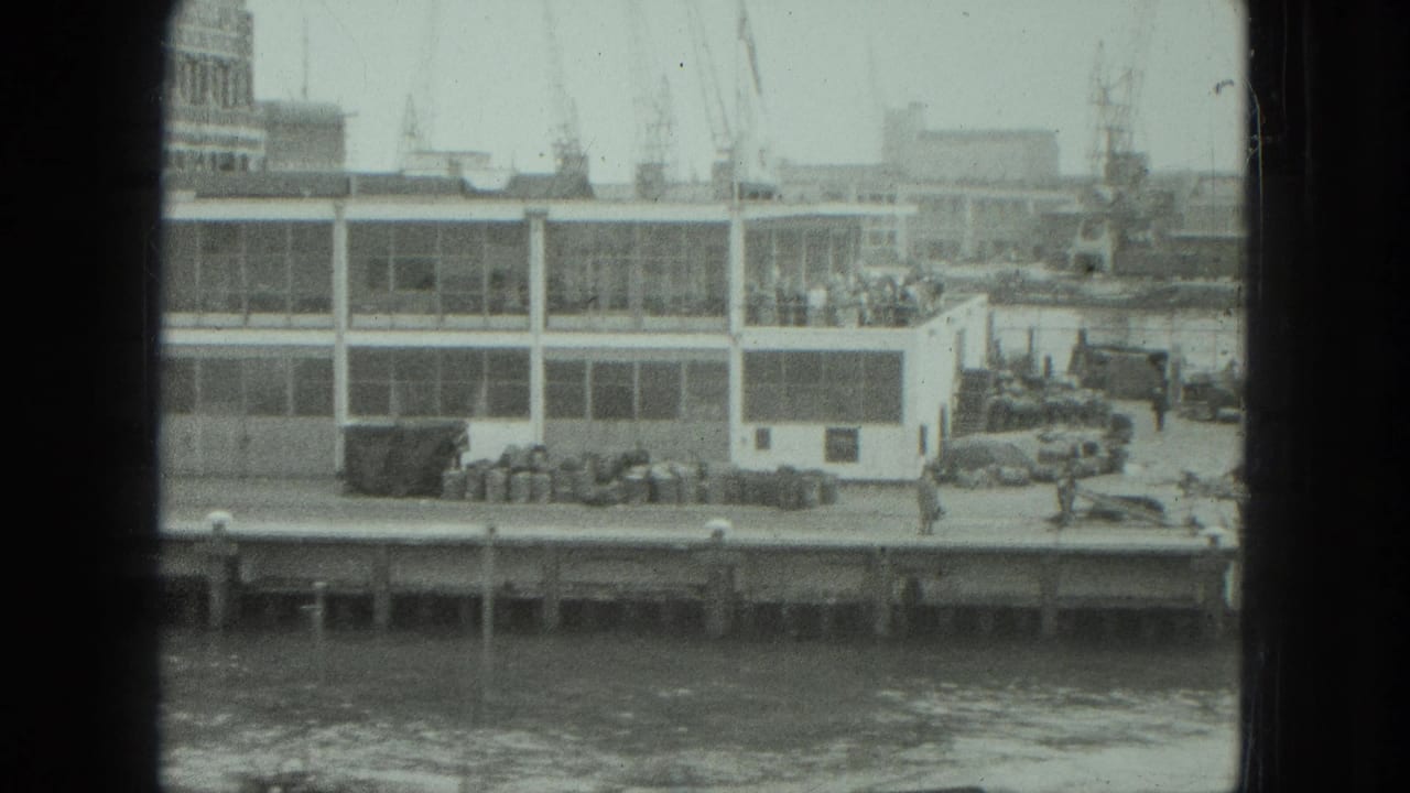 Dockside building, cranes, parked cars, and people along the waterfront.