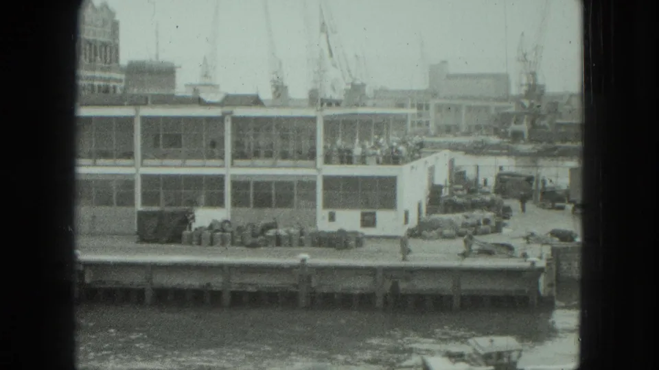 Soft 8mm view of a 1975 Venice dock area, with people near a waterfront building and cranes rising behind it.