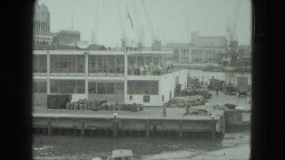 1975 Venice harbor view through a dark frame, showing a dockside crowd, glass-fronted building, cranes, vehicles, and choppy water.