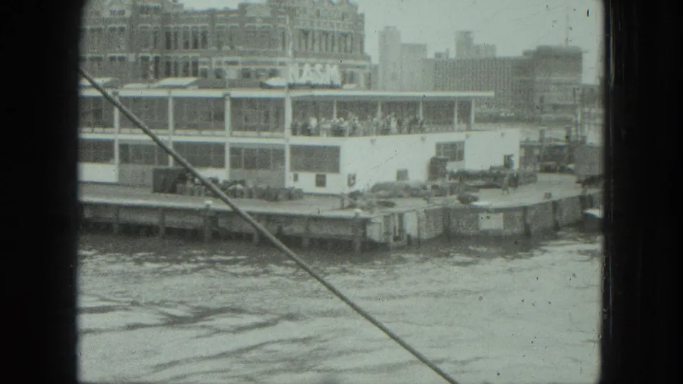 1975 8mm travel frame from a boat, showing a docked waterfront passenger vessel, buildings, water, and a diagonal cable in the foreground.