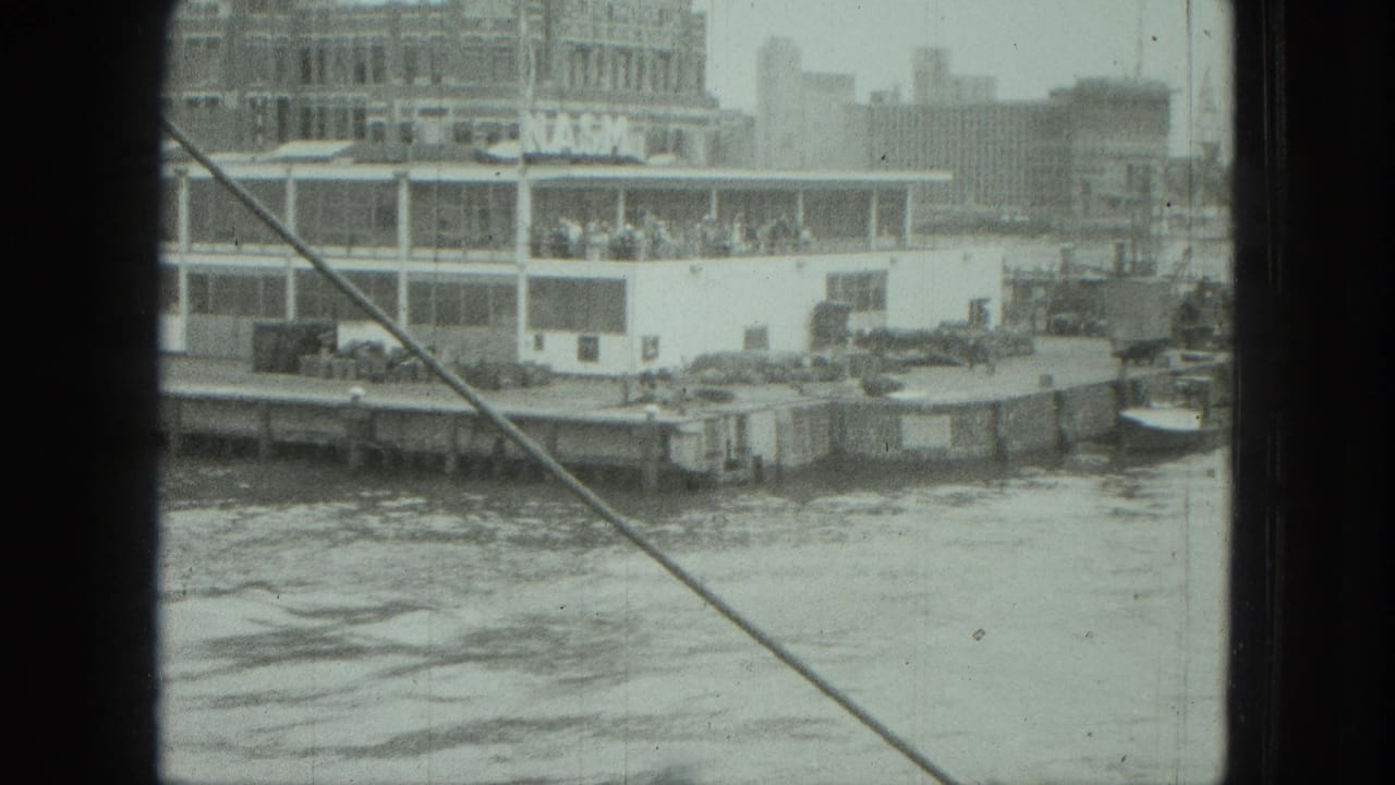 1975 8mm frame of a waterfront passenger boat near buildings, with passengers visible and boat-side framing crossing the view.