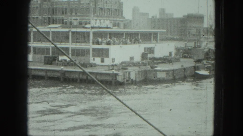 1975 8mm frame of a waterfront passenger boat near buildings, with passengers visible and boat-side framing crossing the view.