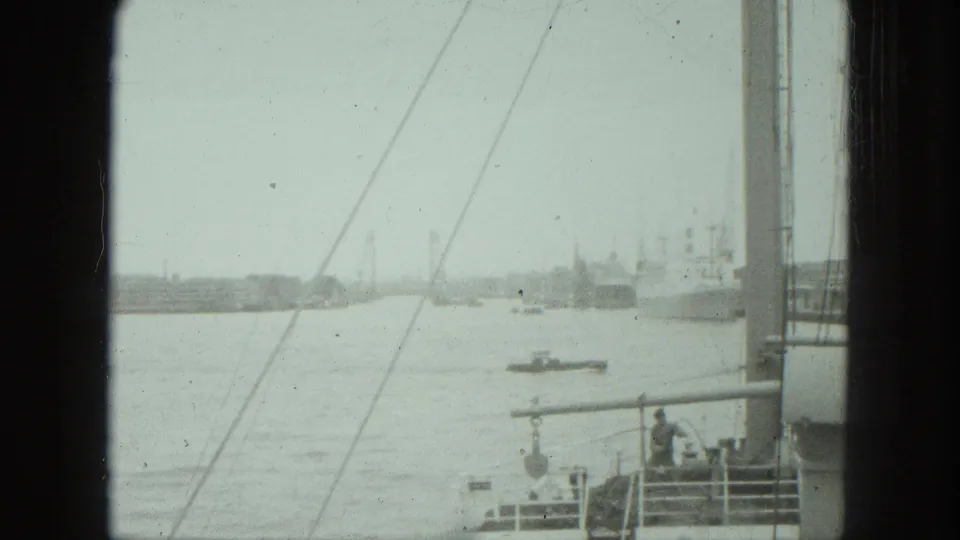 1975 boat-view frame showing a wider harbor scene with distant vessels, rigging lines, deck railings, and a small boat on the water.