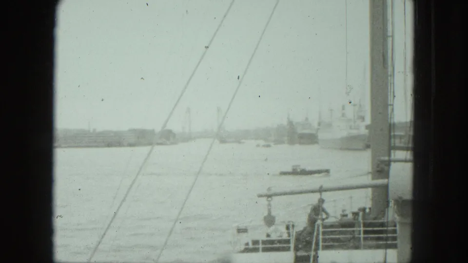 1975 8mm harbor frame from a boat, with a small vessel on the water, ship-side structures, rigging, railings, and a person on deck.