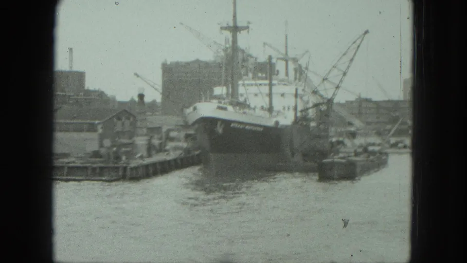 1975 Venice port frame with a cargo vessel at the pier, cranes, small boats, and industrial waterfront structures.