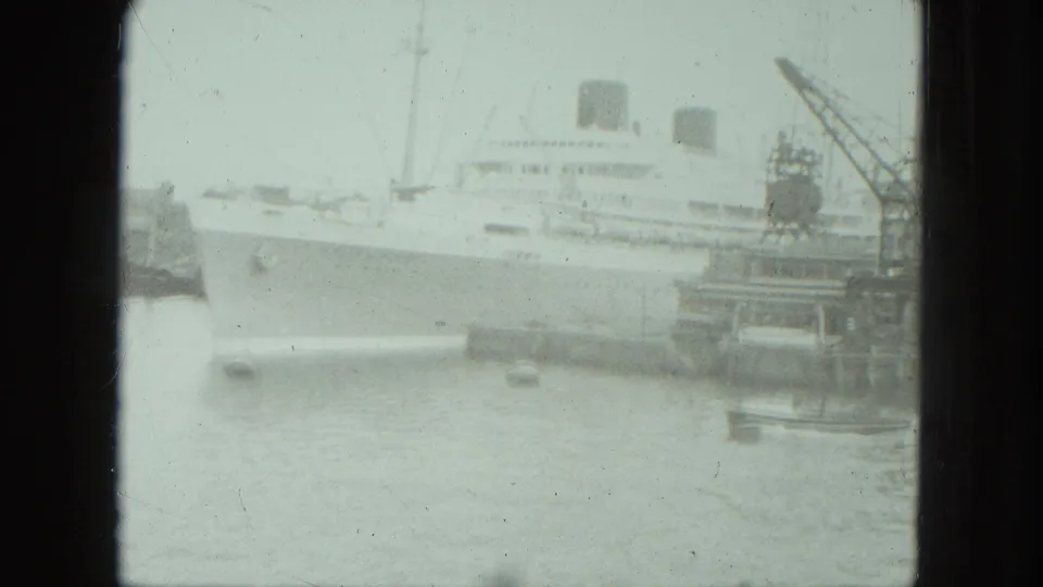 Soft 8mm frame of a large passenger ship at Venice harbor, with cranes and small boats visible across the water.