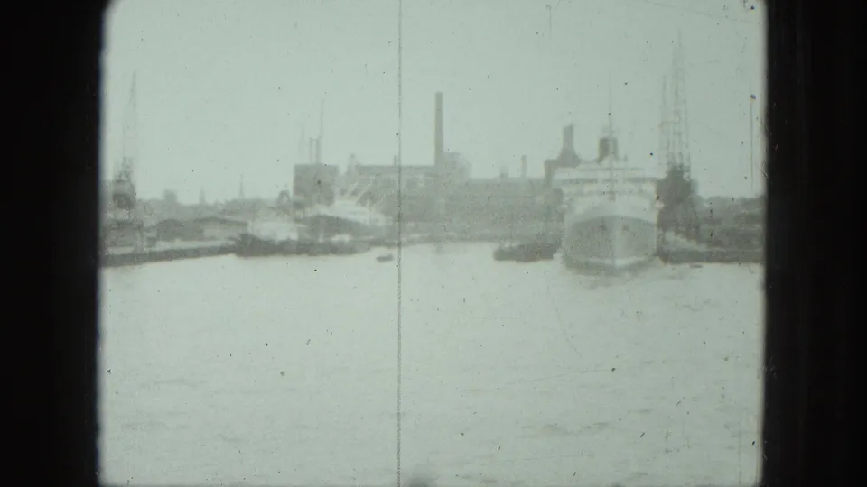 Wider 1975 Venice harbor view with small boats, quay structures, and industrial waterfront forms under pale 8mm exposure.