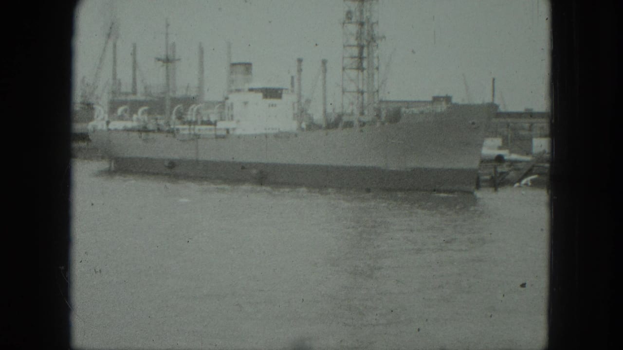 Large ship moored across calm harbor water with cranes and masts behind it.