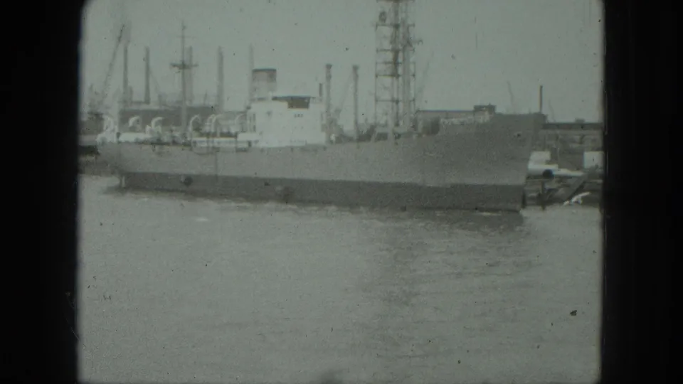 1975 Venice harbor view with a large moored ship, calm water, cranes, masts, and dockside industrial structures in soft 8mm footage.