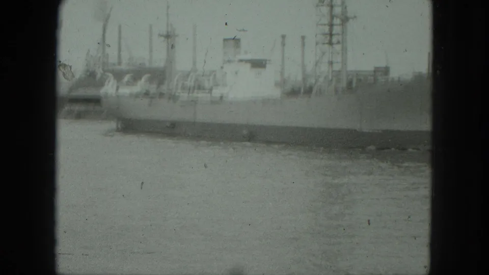 Grainy 8mm frame from Venice in 1975 showing a blurred moored ship, waterfront cranes, masts, and pale harbor water.