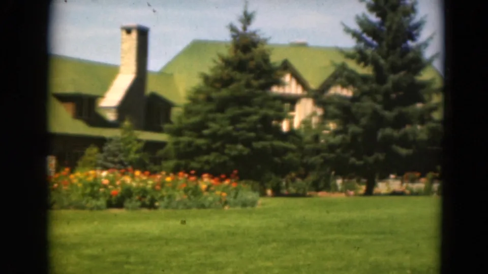 Rustic building with green roof and stone chimney seen across a broad lawn with flower beds and evergreens.