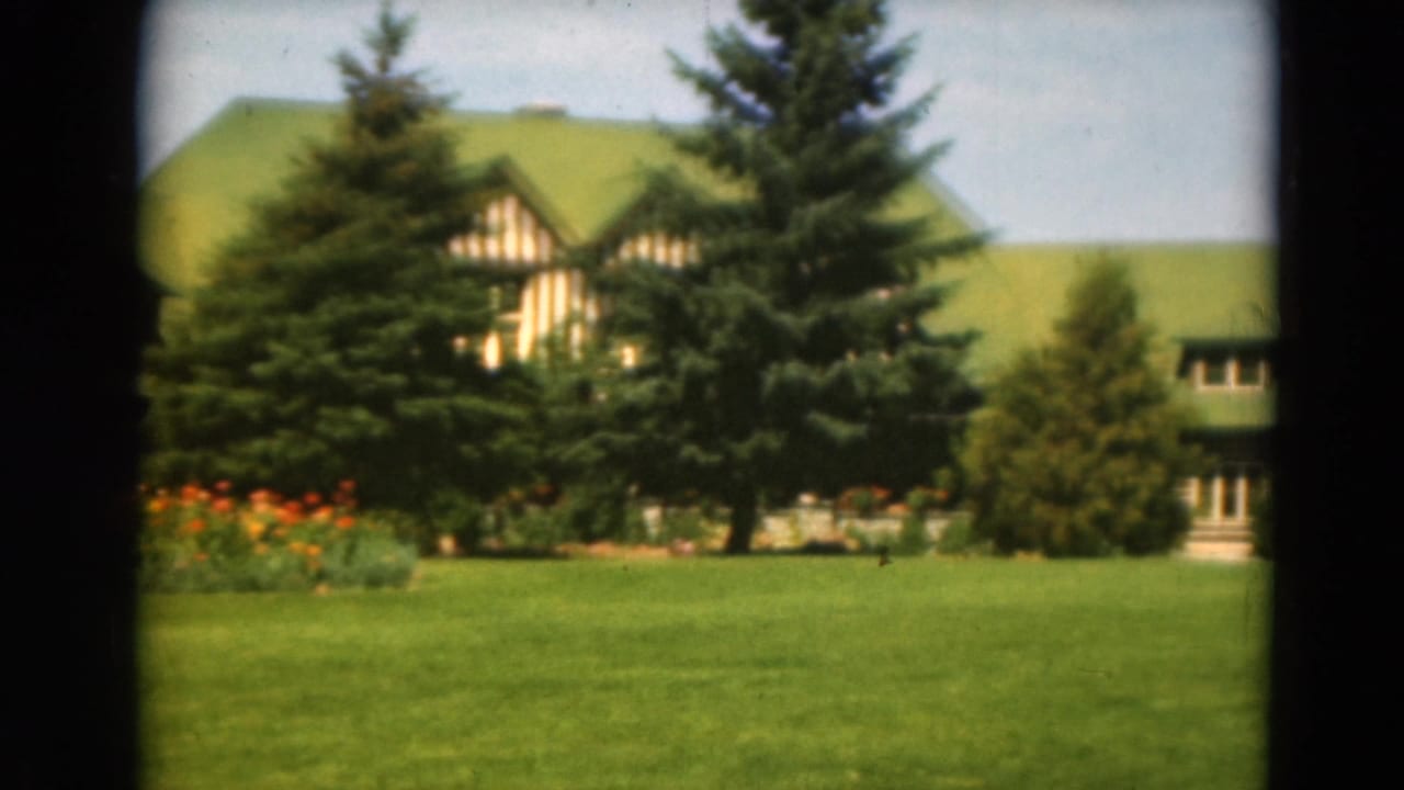 A landscaped lawn and flower bed sit before evergreens that partially hide a green-roofed rustic building.