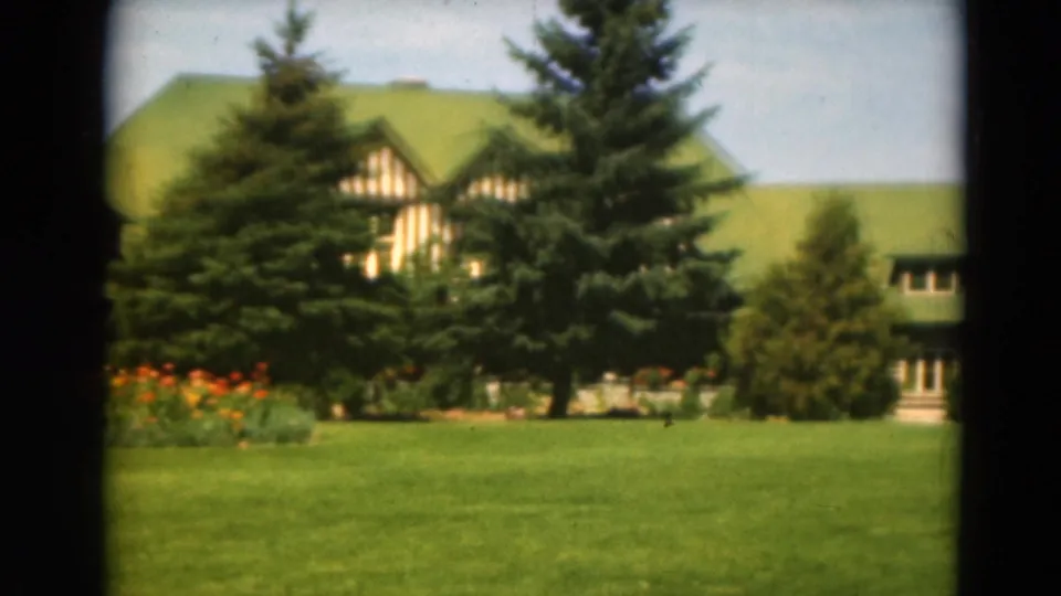 A landscaped lawn and flower bed sit before evergreens that partially hide a green-roofed rustic building.