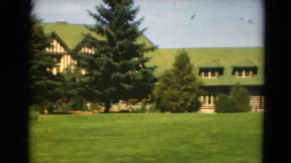 The pan reveals more of the long green-roofed structure with dormers, lawn, evergreen trees, and flowering plants.