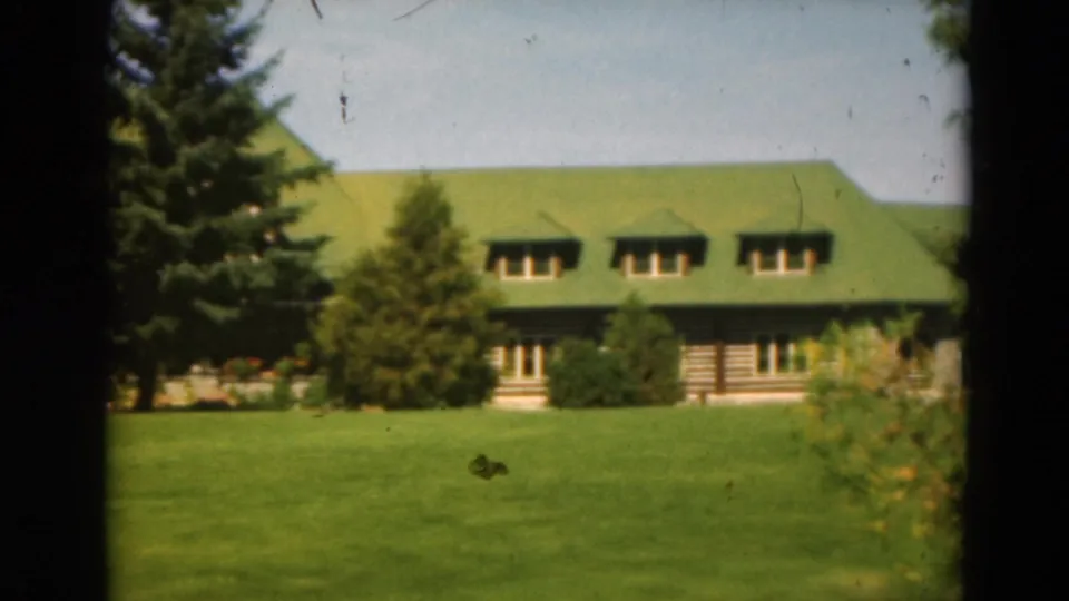 A long rustic exterior with green roof and dormers stands behind a wide lawn, evergreens, shrubs, and flowers.