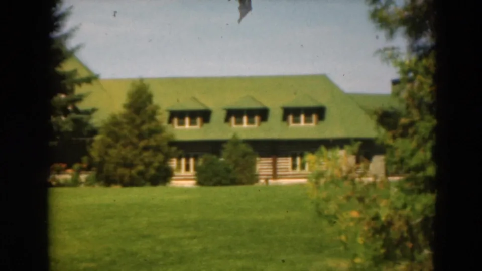 1975 Duluth home-movie frame showing a log-style building with a green roof, dormer windows, lawn, shrubs, and trees.