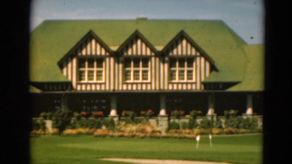 1975 Duluth frame of a Tudor-style building with striped gables, a green roof, large windows, porch, lawn, and flower beds.