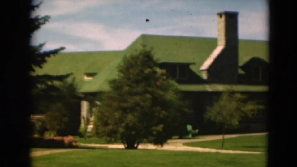 1975 Duluth home-movie frame showing a lodge-like building with a green roof, stone chimney, trees, lawn, and curved path.