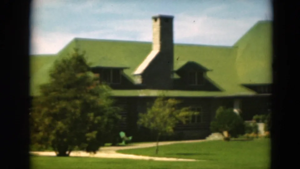 1975 Duluth frame showing a green-roofed lodge or house with a stone chimney, shaded trees, lawn, walkway, and shrubs.