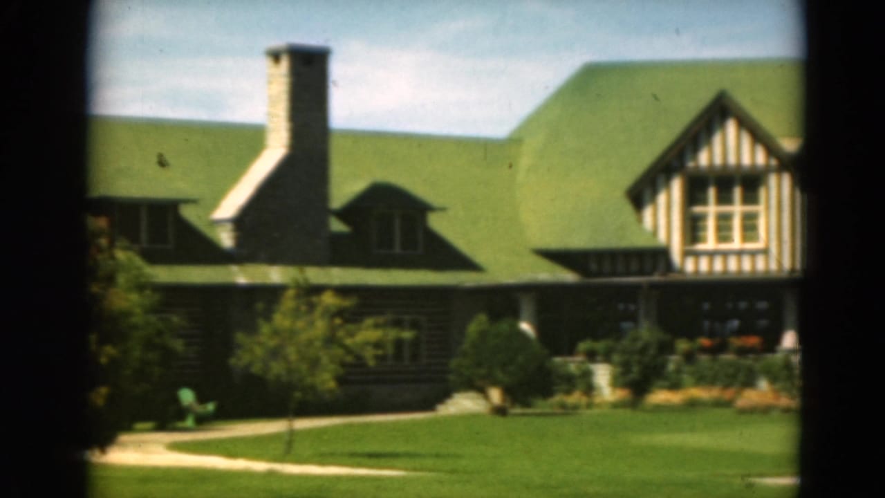 Green-roofed timber-framed building with stone chimney, lawn, path, and dark foreground framing.