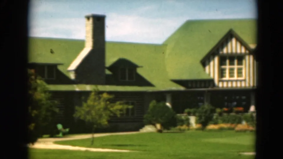 1975 Duluth home-movie frame showing a green-roofed timber-framed building, stone chimney, porch, lawn, and garden through dark foreground framing.