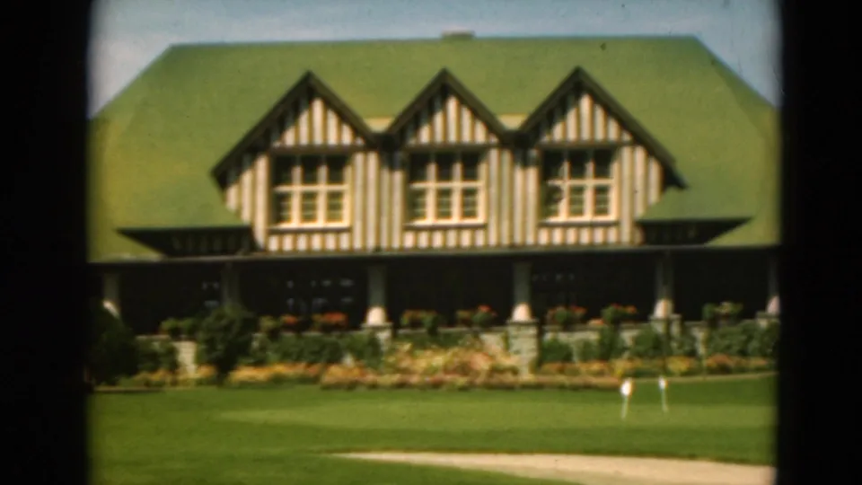 1975 Duluth home-movie frame centered on three striped timber gables, a green roof, shaded porch columns, flowerbeds, and open lawn.