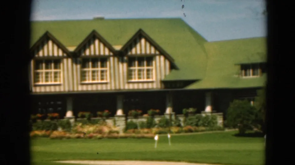1975 Duluth frame showing timber-framed gables beneath a green roof, with porch columns, planted beds, lawn markers, and dark foreground framing.