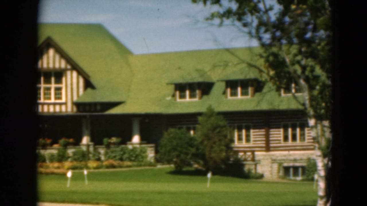 Green-roofed building with Tudor gable, log-style wall section, porch, lawn, and trees.