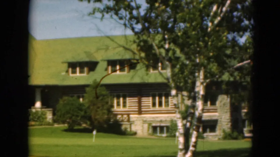 1975 Duluth 8mm frame showing log-style wall details, a stone foundation, green roof, birch trees, and open lawn.