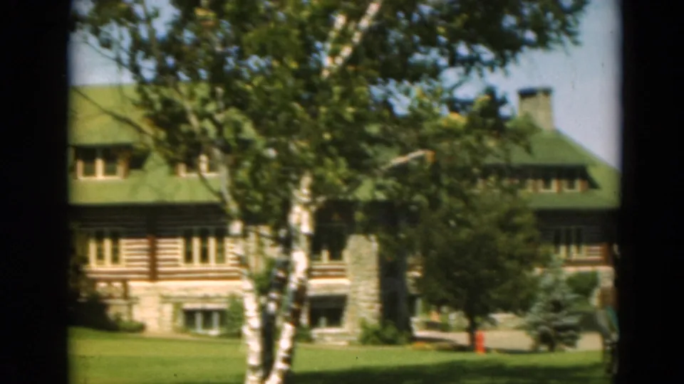 1975 Duluth frame of the house partly screened by mature trees, with green roof, log-style siding, stone base, and lawn.