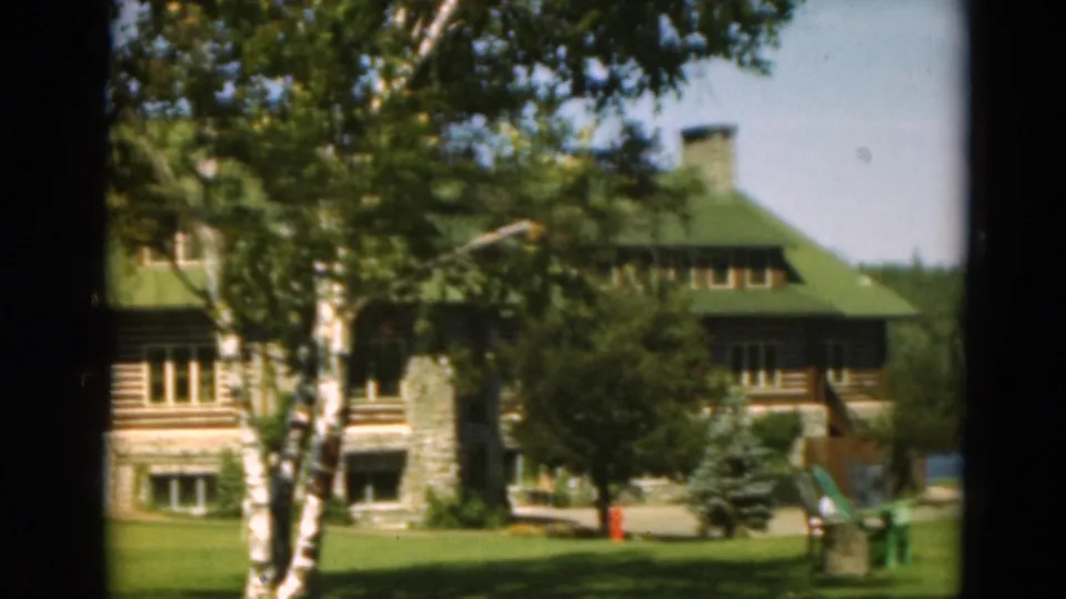 1975 Duluth-area home-movie frame showing a log lodge with a green roof, stone base, lawn, and trees near water.