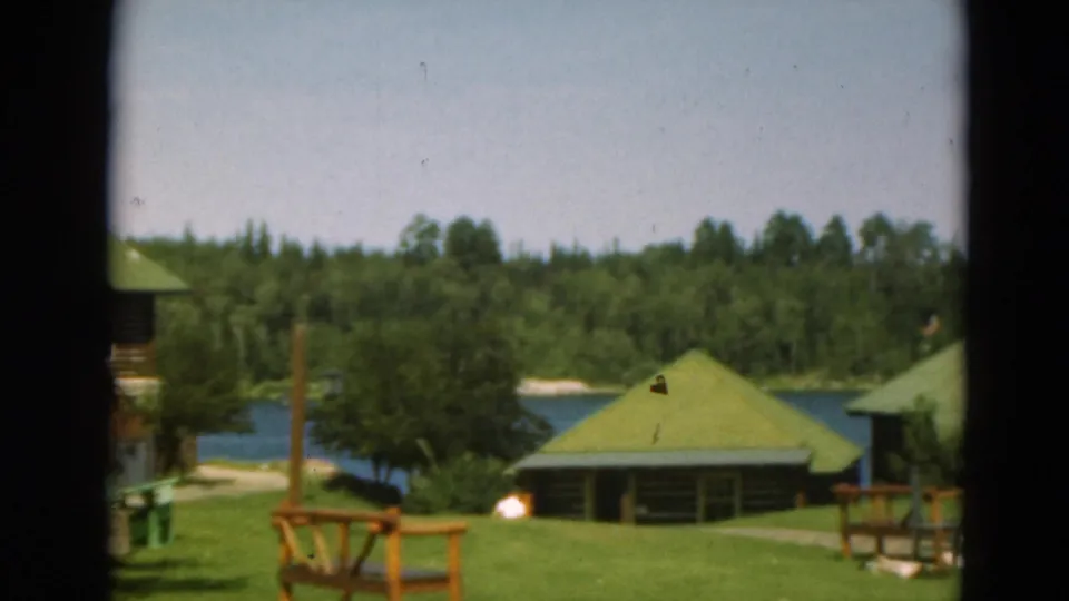 1975 Duluth lakeside resort view with a small green-roofed cabin, lawn, railings, water, and wooded background.