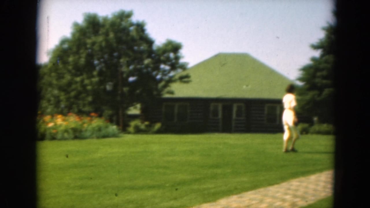 Woman in a pale dress walking across a green lawn near a log cabin and trees.