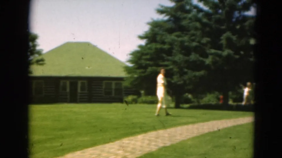 Woman in a light dress walks across resort-like grounds in Duluth, Minnesota, with a green-roofed cabin behind her.