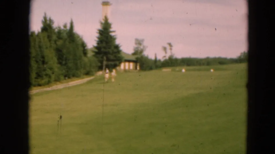 Wide 1975 home-movie view of open green grounds near Duluth, with small walkers, trees, a cabin, and a distant tower.