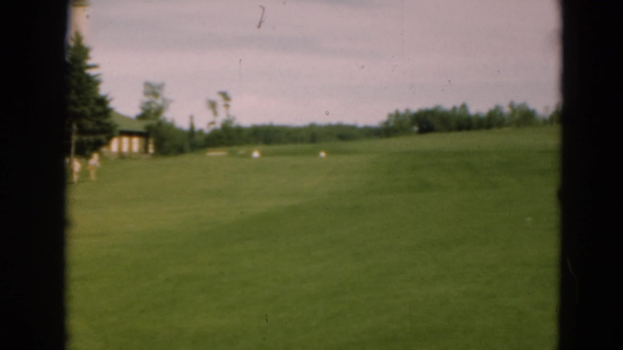 Green golf fairway seen through dark film-frame edges with trees and a small structure in the distance.