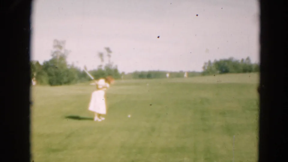 A woman golfer in a light dress appears on a Duluth, Minnesota fairway in 1975 home-movie footage, with trees and open grass behind her.