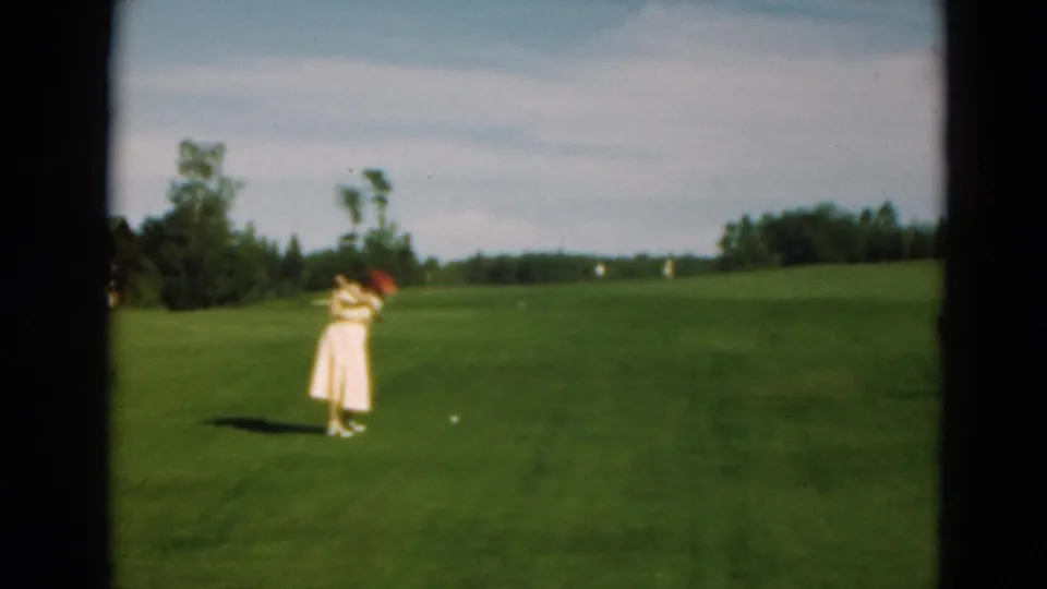 1975 Duluth home-movie frame of a woman in a light dress and red hat on a green golf fairway, with a golf ball visible nearby.