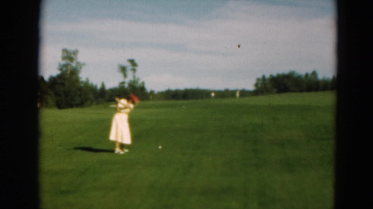 Woman in a light dress and red hat prepares to swing a golf club on a green fairway.