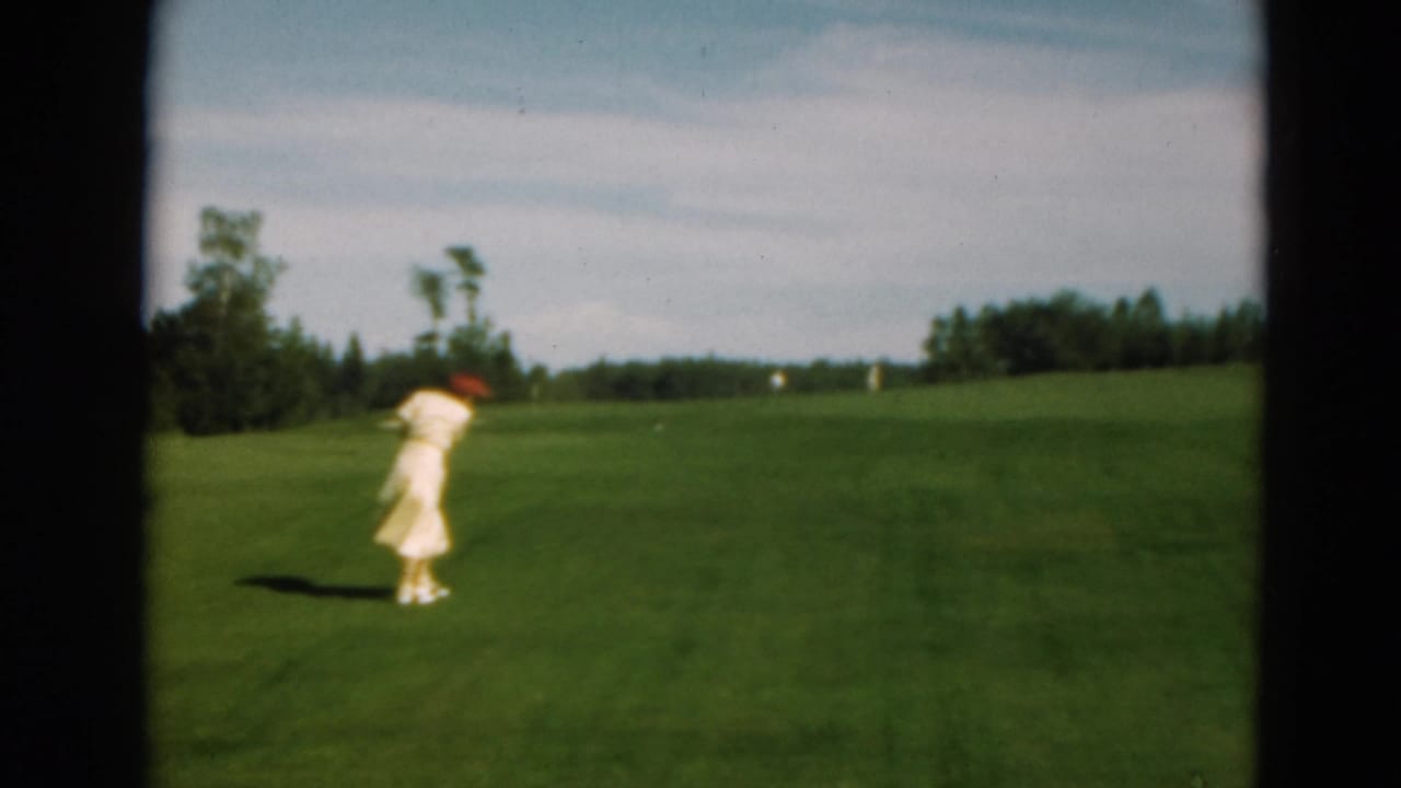 A 1975 Duluth home-movie frame shows the woman following through after a golf swing on an open course.
