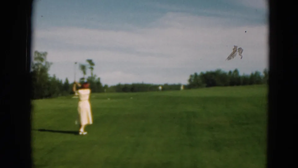 A woman golfer in 1975 Duluth stands on a green fairway with her club raised, framed by trees and sky.