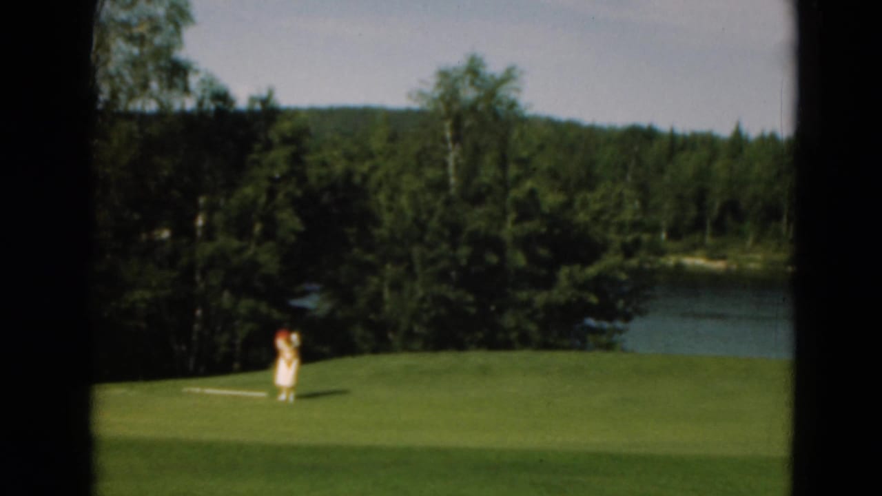 Distant golfer in a light dress stands on a green with trees and a lake behind her.