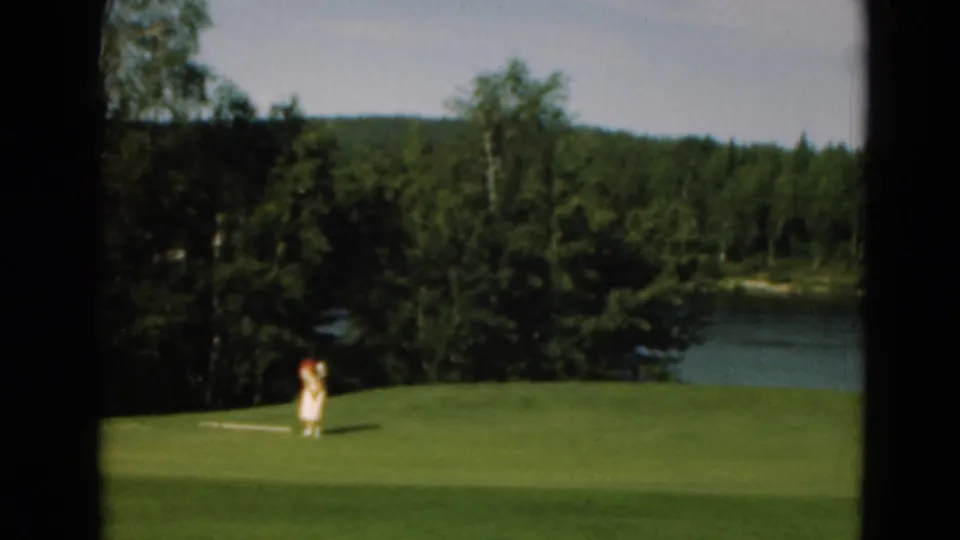 In supported 1975 Duluth, Minnesota footage, a distant woman in light clothing stands on a golf green beside a wooded lake.