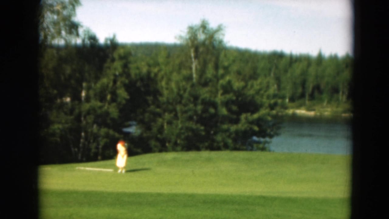 Golfer swings on a green near a lake bordered by dense trees.