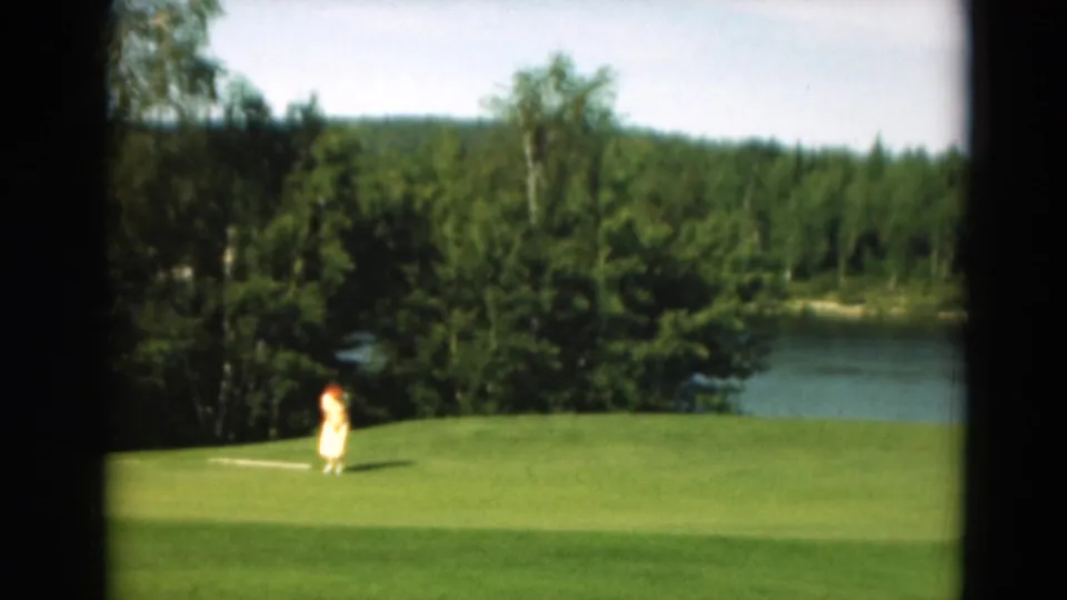 A distant woman golfer swings on a green in the supported Duluth, Minnesota 1975 home-movie scene.