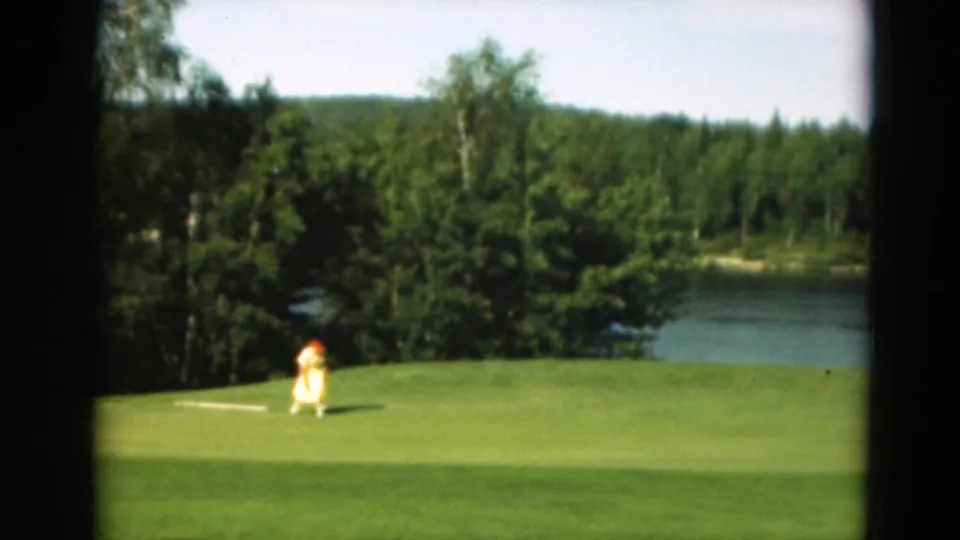 The distant golfer bends or follows through after a shot on a lakeside green in supported 1975 Duluth footage.